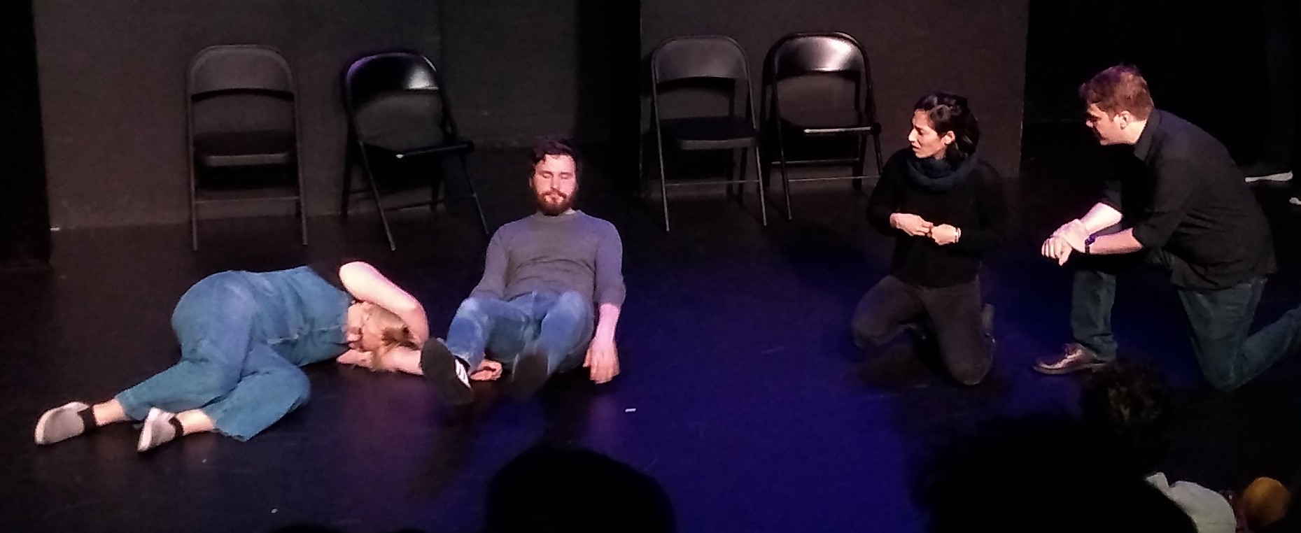 Stage right: two people lie on the floor in contorted poses. Stage left: a woman kneels on the floor looking at them, while a man bends down on one knee to speak to her.