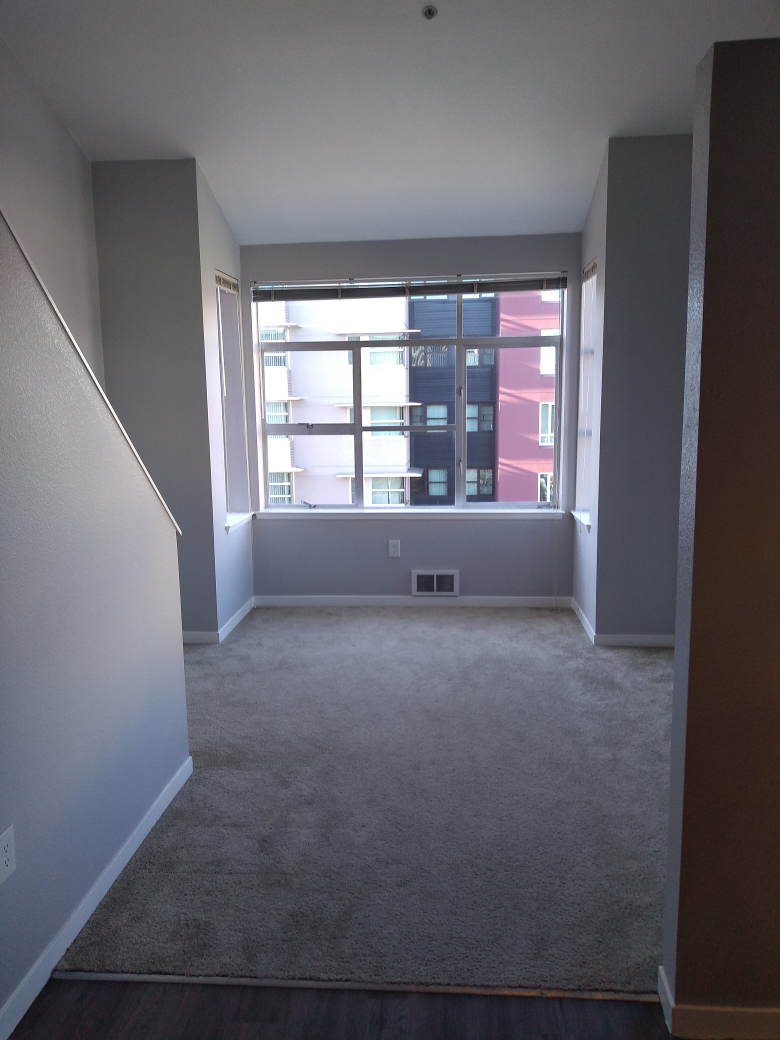 View of an empty carpeted room, with a large picture window overlooking some buildings across the street.