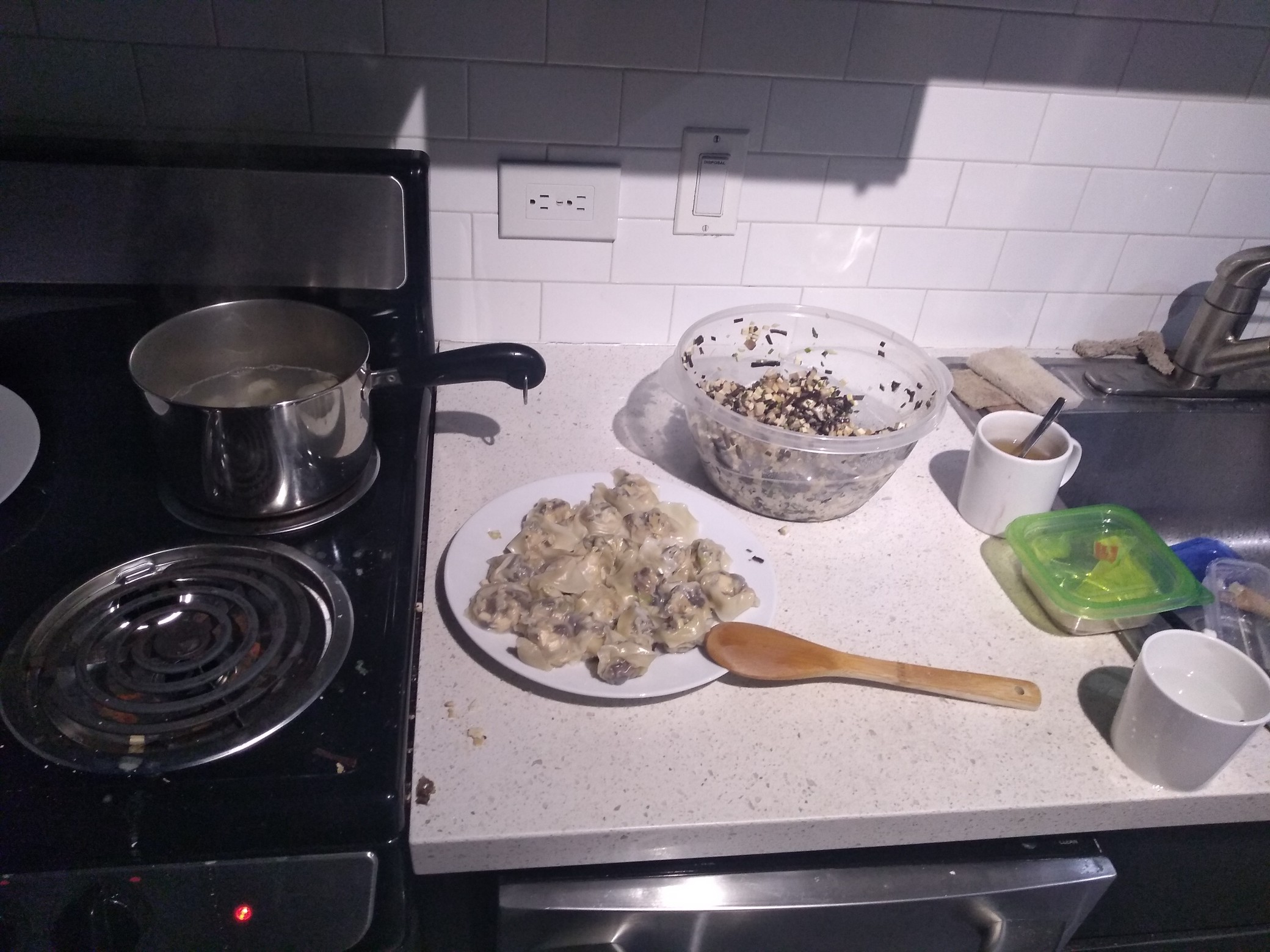 A plate full of slightly lumpy dumplings, surrounded by various accoutrements of cooking including a boiling pot of water and a bowl full of chopped mushroom stuffing.