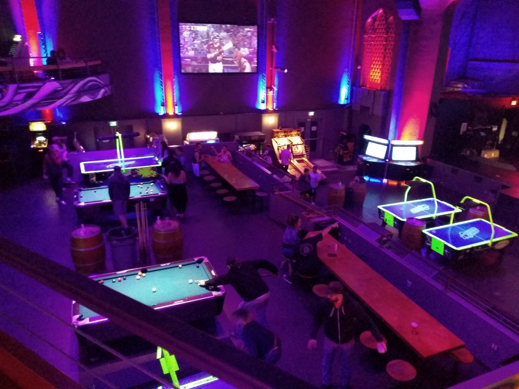 A large, dim room lit in neon colors, with various pool tables, air hockey tables, and arcade machines scattered about.