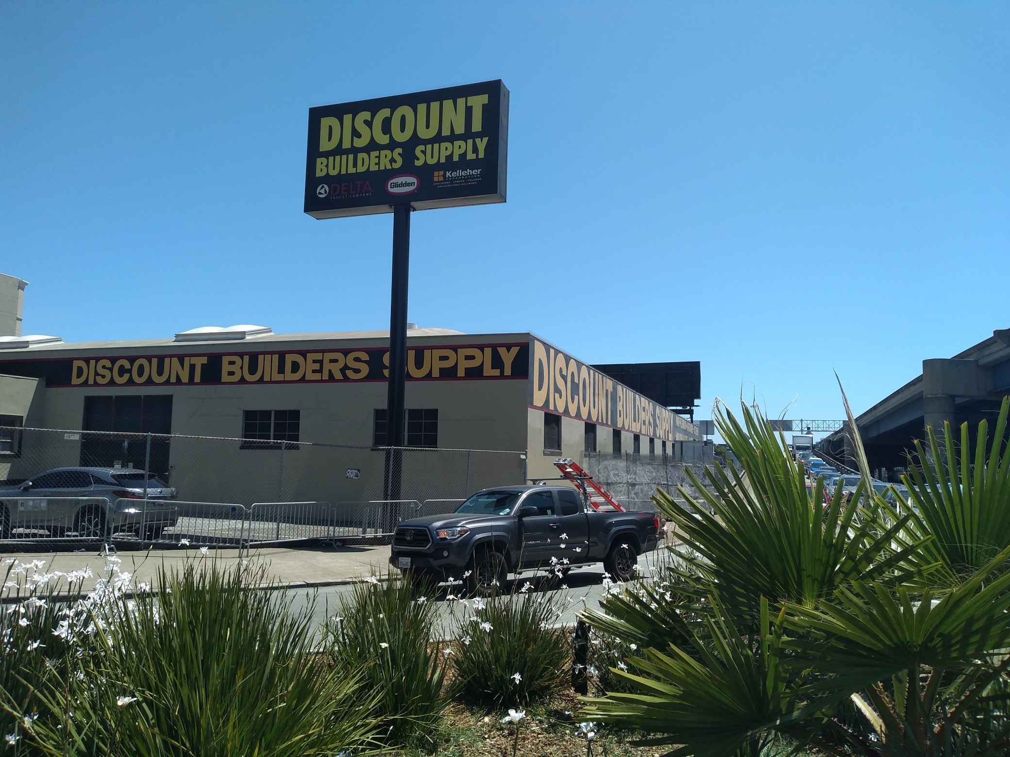 A large single-story building next to a freeway, with “DISCOUNT BUILDERS SUPPLY” plastered all over it in large yellow-on-black letters.