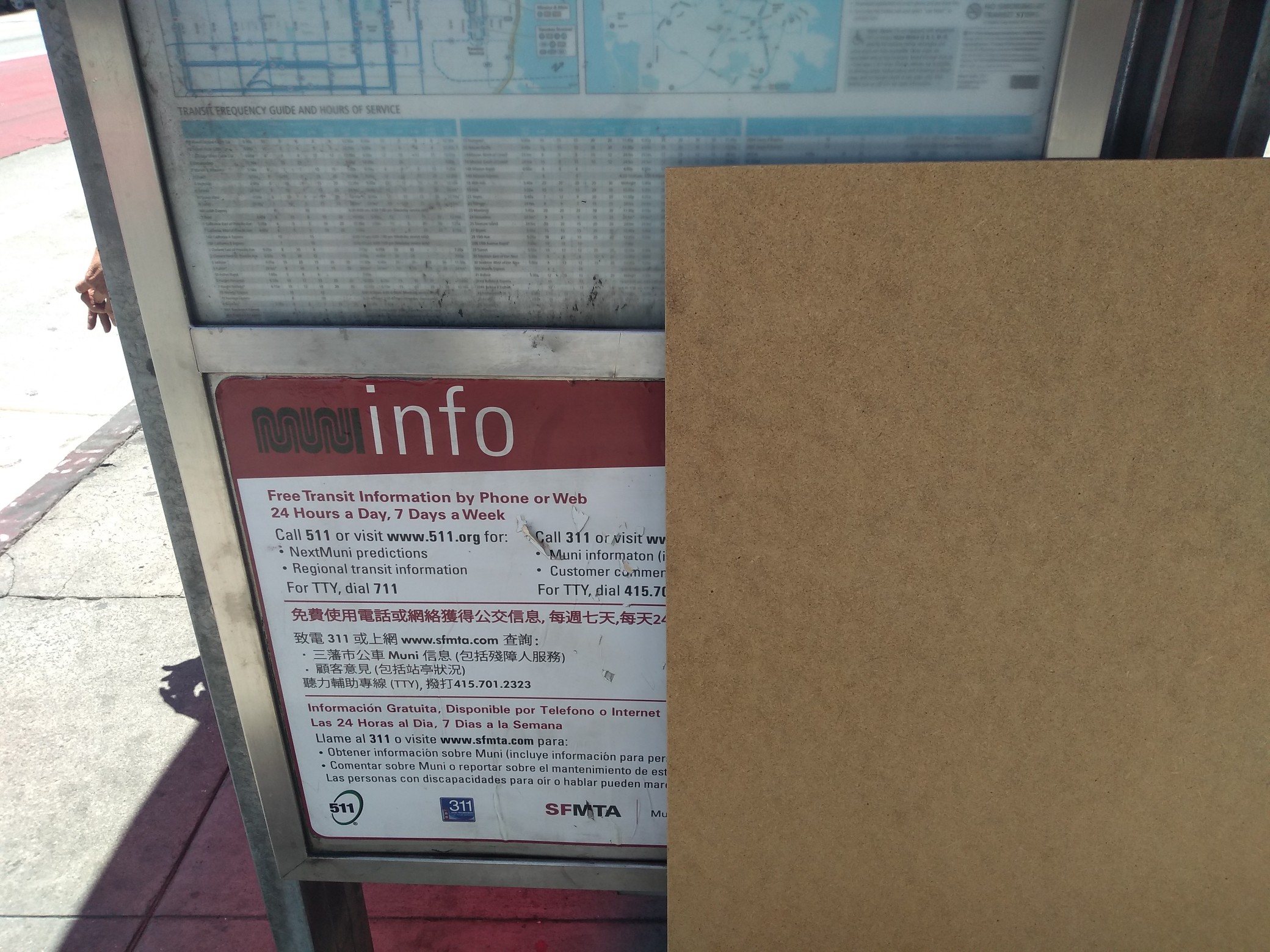 A large piece of brown-speckled hardboard leaning up against a Muni bus shelter.