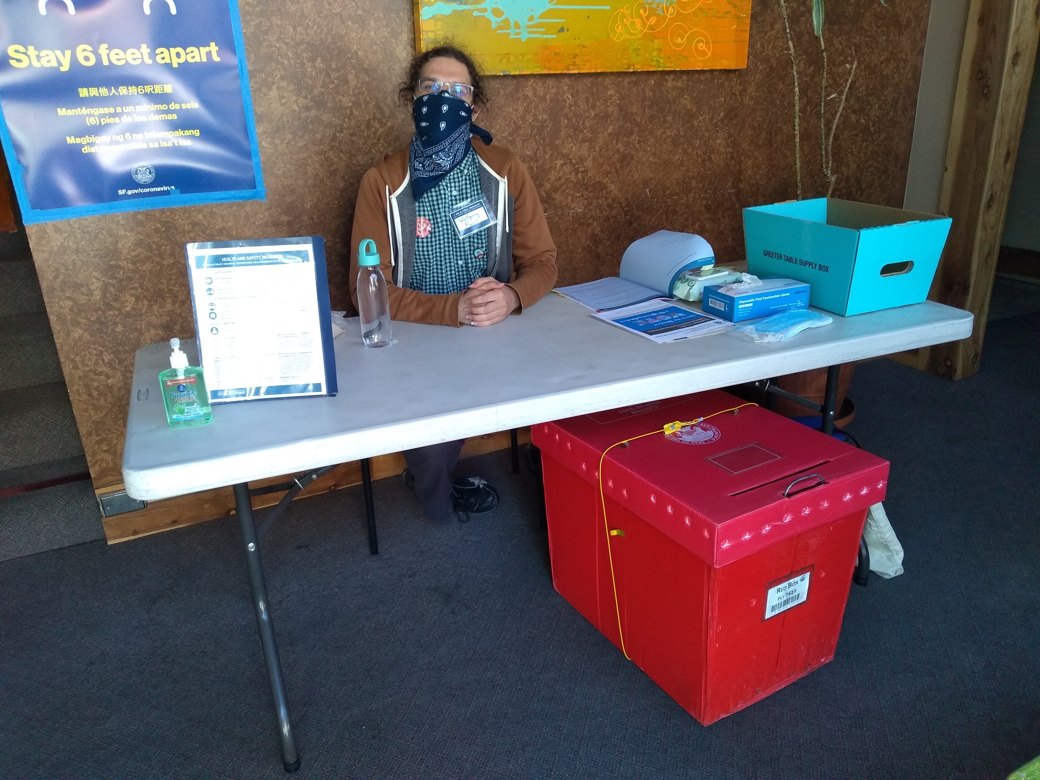 Wolf sits at a folding table, surrounded by various posters, safeguarding a large red box for vote-by-mail ballots.