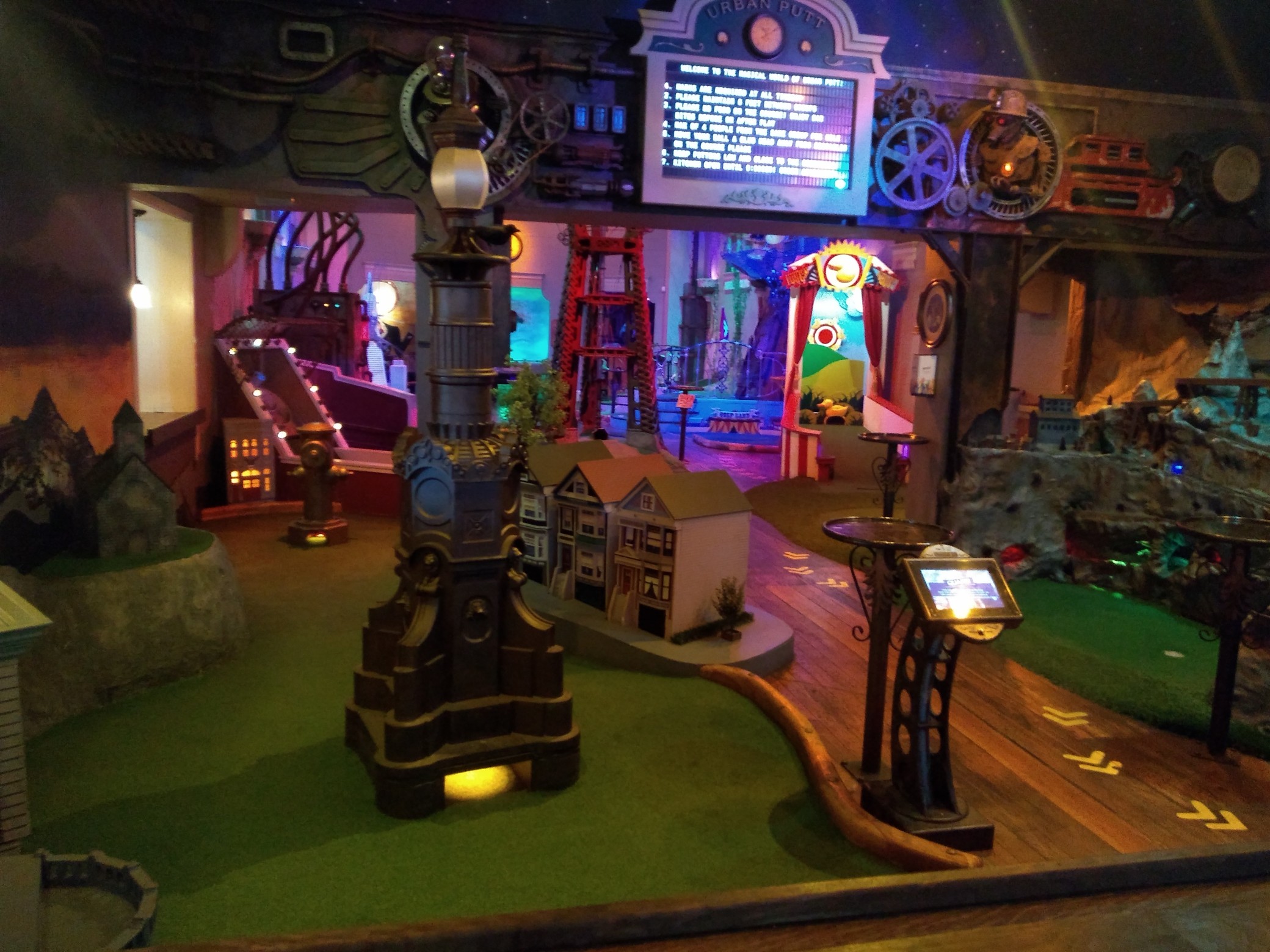 A room with several mini-golf holes. In the foreground is a green surrounded by model houses, with a large lamppost in the center. To the right is a large model of a mountain with gold mines, and further back can be seen a Duck Hunt-themed hole and some more indistinct scenes. Overhead is a large display of exposed gears and pipes.