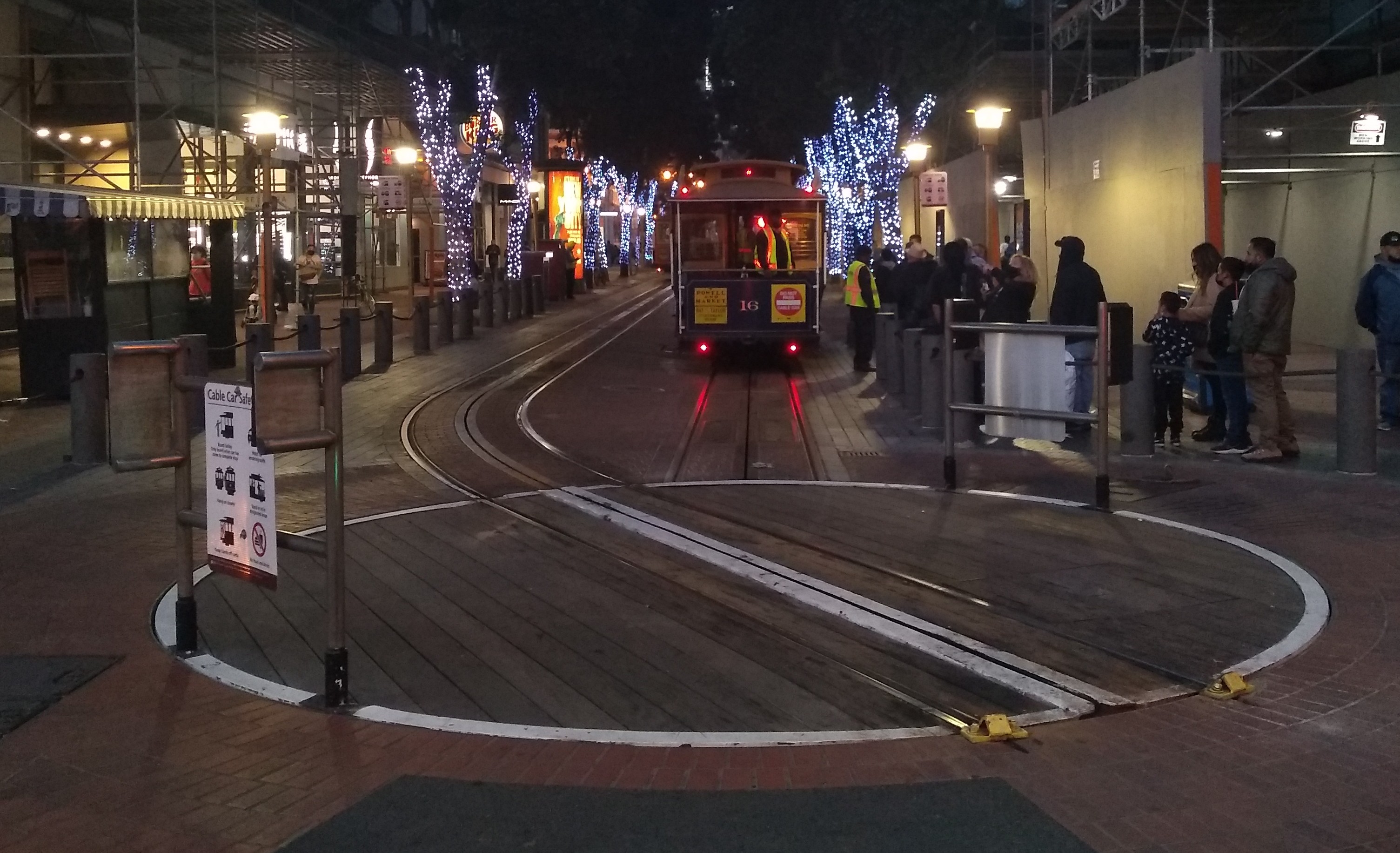 Dusk. In the foreground, a railroad turntable, with a cable car just past it. In the background, tree trunks wrapped in Christmas lights.